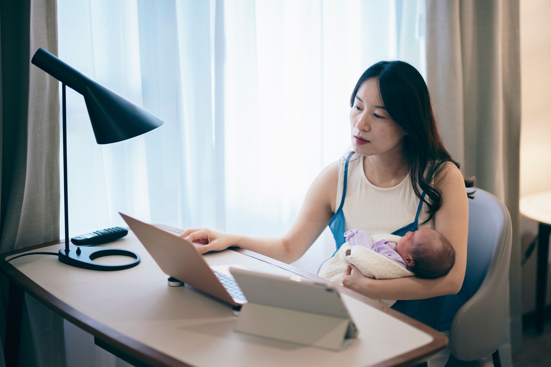An Asian mother takes care of her baby while managing work tasks and household finances on her laptop.She balances the responsibilities of work and family, embodying the concept of balancing work and childcare.