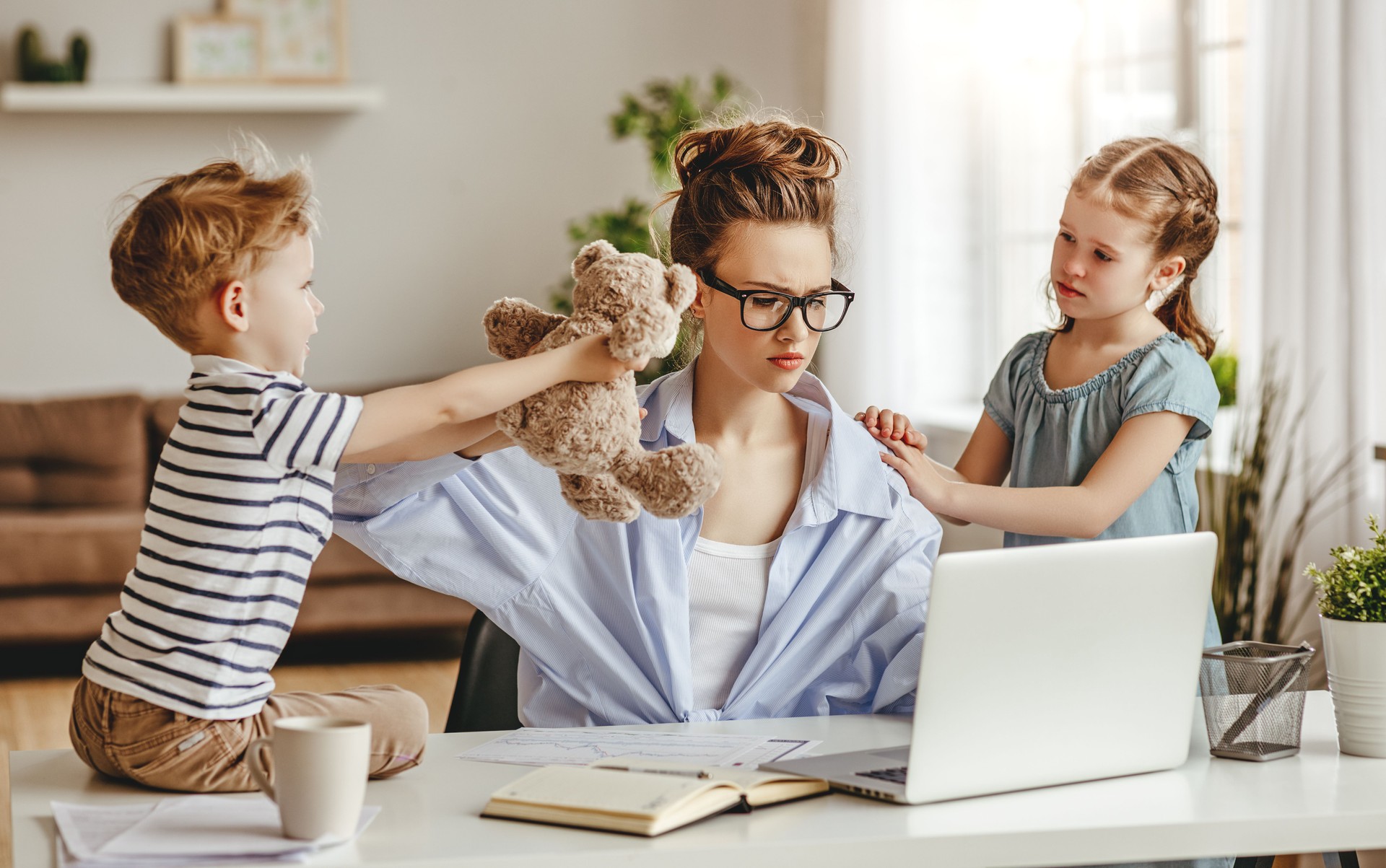 Little children distracting dedicated young woman working on laptop at home